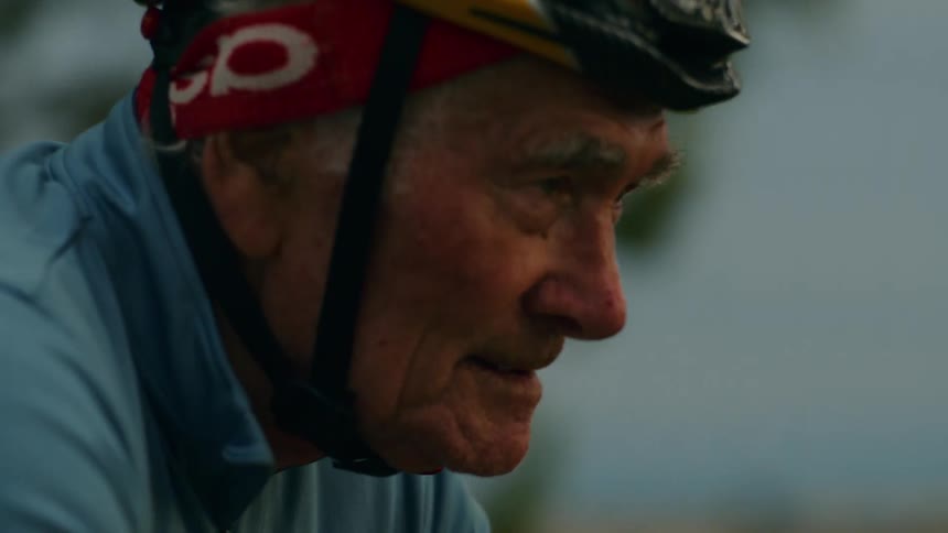 A close up shot shows the side profile of an elderly man with white hair wearing a cycling helmet and a light blue jersey