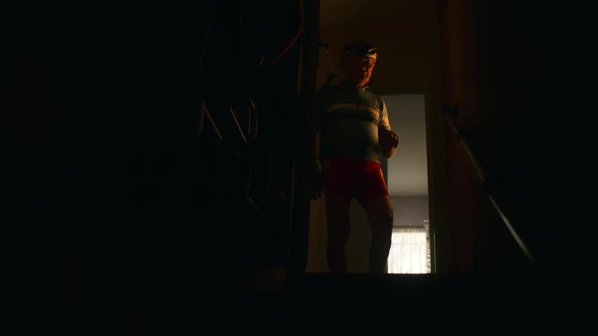 An older man flips on a light at the top of a dark staircase puts on his cycling helmet and then walks resolutely down the stairs into the shadows below