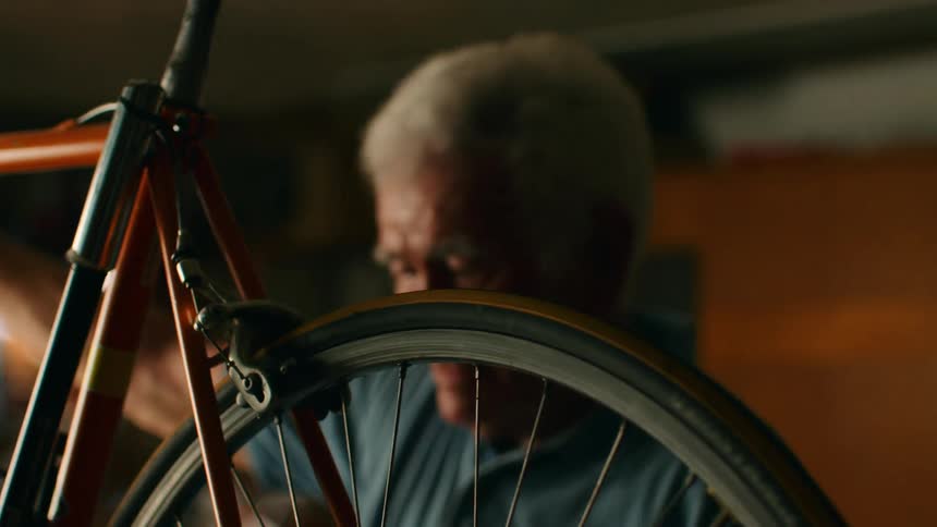 An elderly man repairs a vintage orange bicycle in a dim indoor workshop