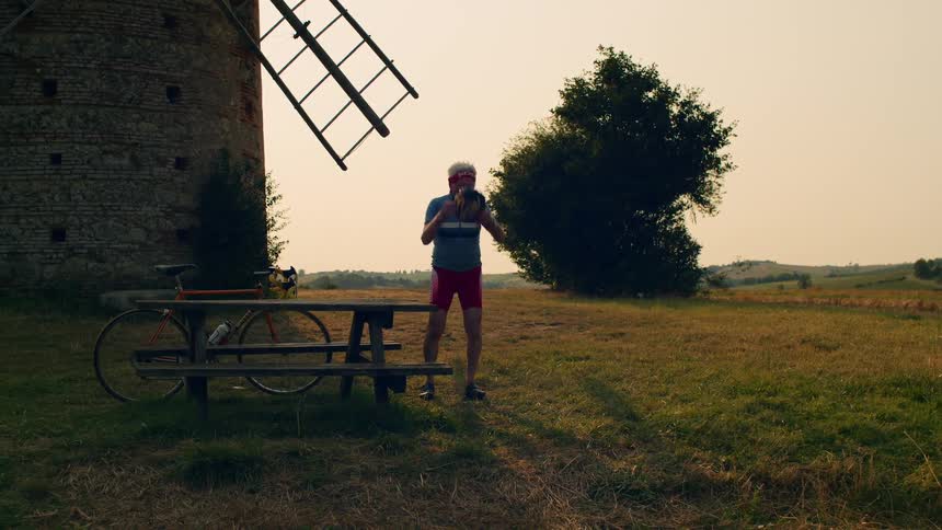 Man walks his bicycle near a building and leans it against a picnic table then takes off his helmet and sits down