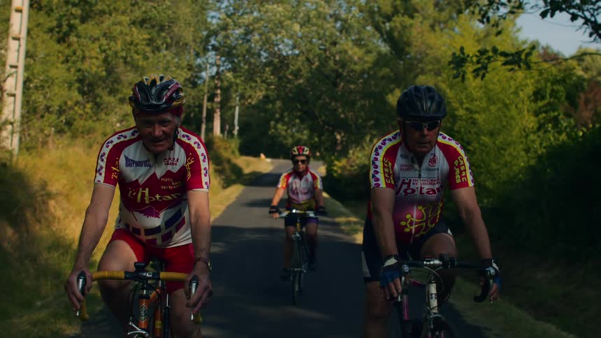 Four male cyclists in colorful jerseys ride along a paved path through a sundappled forest looking forward with steady determination