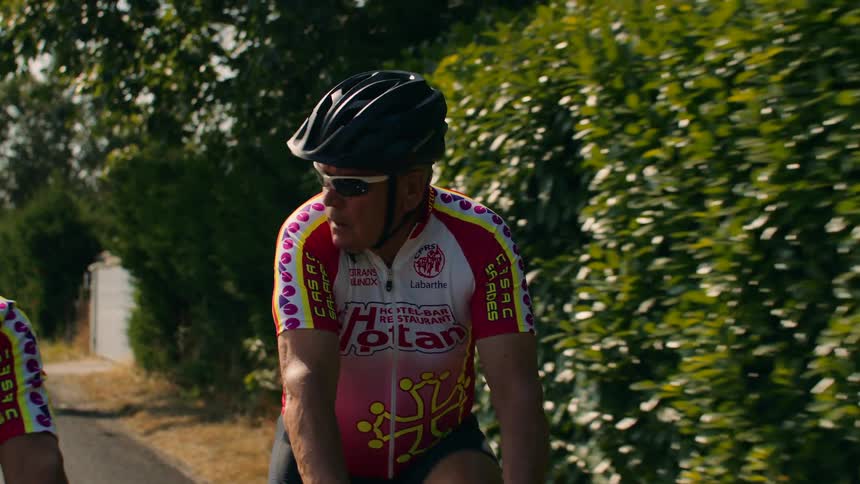 A medium shot shows a group of men in cycling jersey and helmet riding a bicycle along a road lined with green bushes during the day