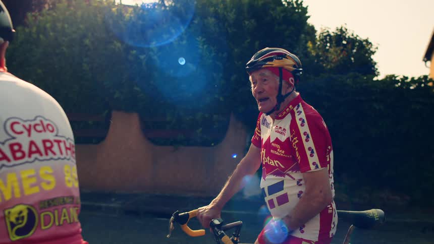 Cyclists in red and white uniforms riding on sunny road with greenery and buildings in the background