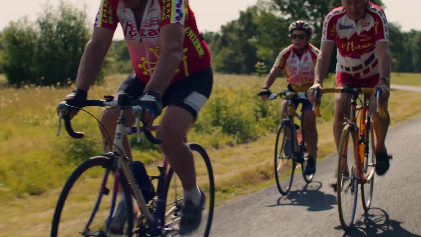 Cyclists in red yellow and white uniforms riding on rural road bordered by fields and trees