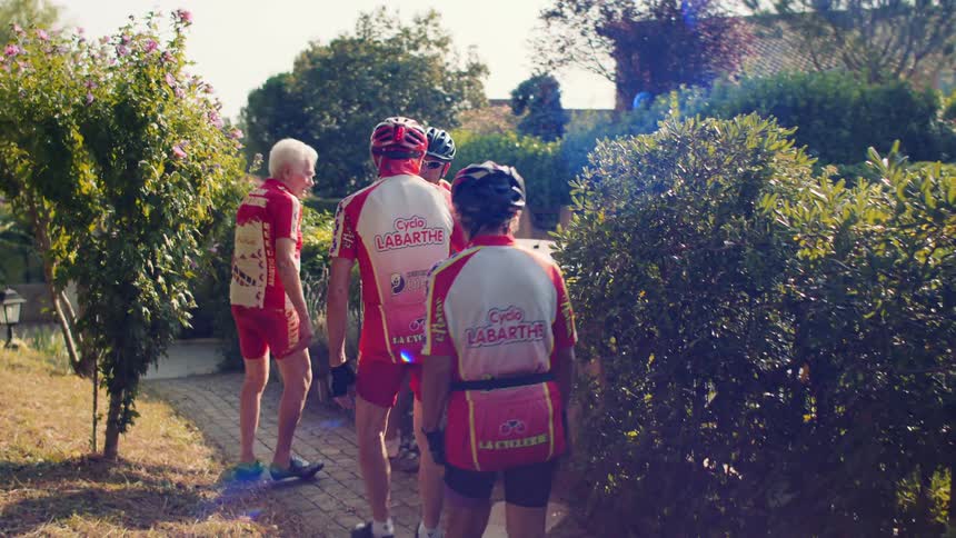 Cyclists walking with bikes along narrow path bordered by flowering bushes and greenery on sunny day
