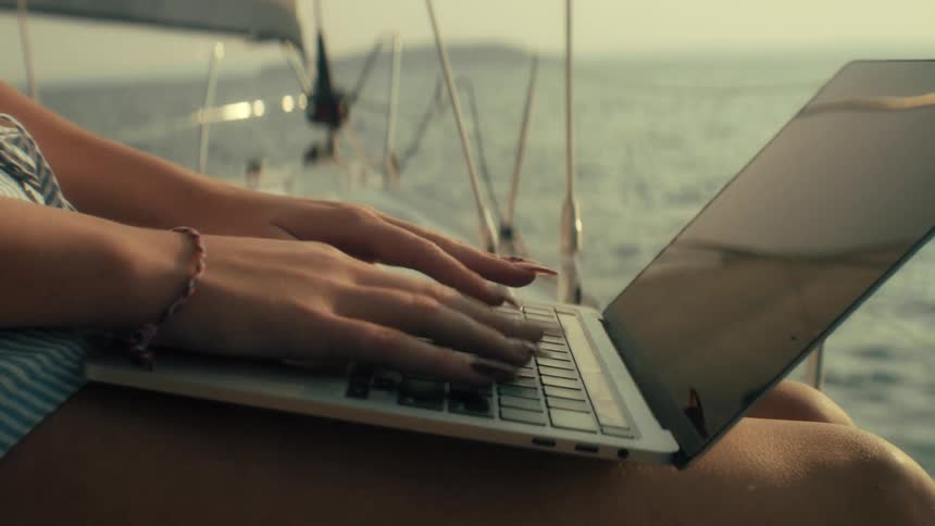 Person typing on laptop on lap with ocean view from sailboat at sunset