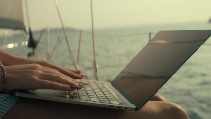 A close up shot shows a woman on a boat opening a laptop and beginning to type while on the water at sunset