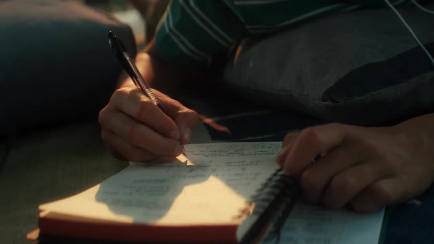 A close up shot shows a young boy smiling and wearing headphones while lying down and writing in a notebook outside during sunset