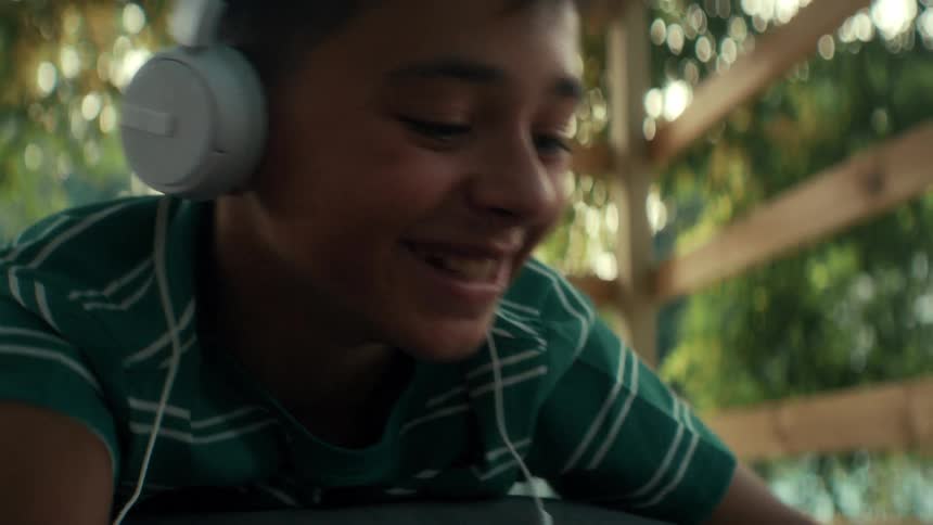 Young boy wearing headphones smiles and laughs while chatting with friends in a virtual classroom in a tree fort
