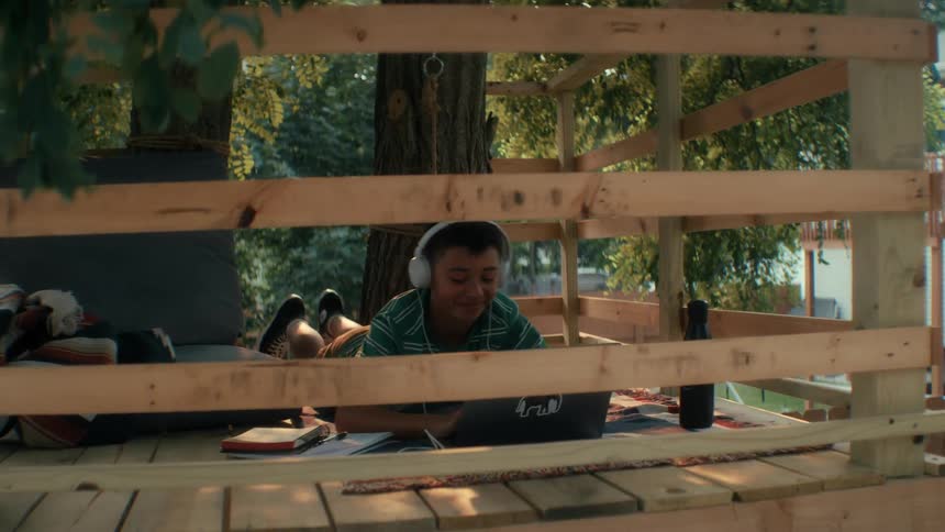 A boy with headphones on smiles while doing homework outside in his tree fort during the summer