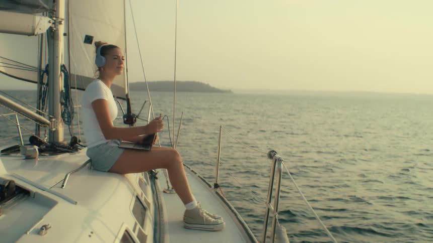 Teenage girl sitting on a boat sailing off the coast as she holds a laptop computer