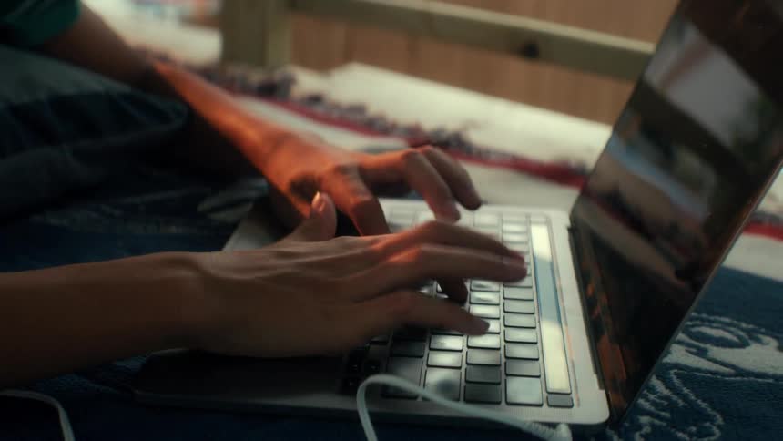 A close up shot shows a persons hands typing on a laptop while they are lying down outdoors during sunset
