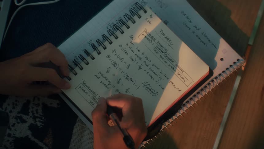 A boys hands are captured in a closeup shot as he writes detailed mathematical notes into a spiral notebook using a blue pen on a wooden desk
