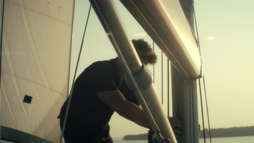 A young man stands on his sailboat at sunset wearing sunglasses as he ties down the sail while warm light glows across the water