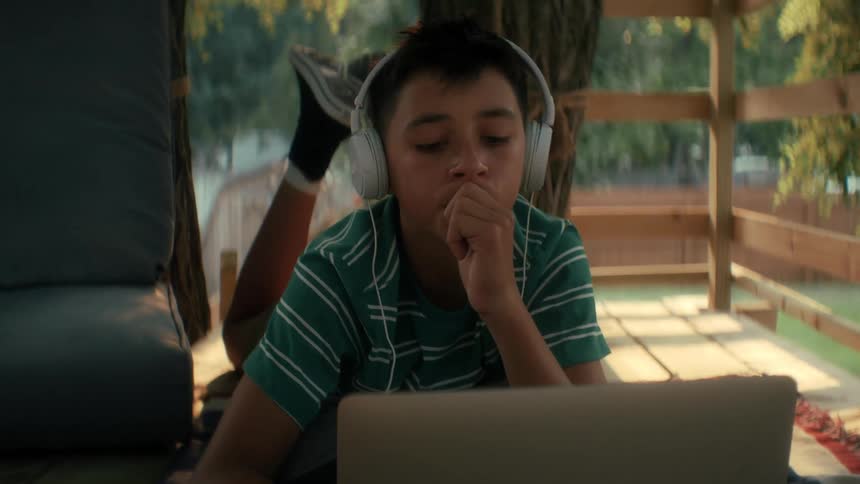 A young boy wearing white headphones lies in his treehouse while looking at a laptop
