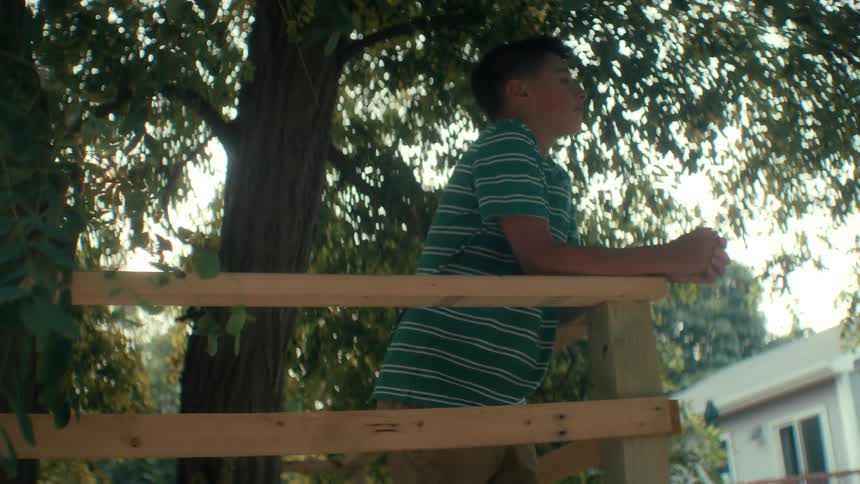A teenager out in nature leans on against railing of he tree fort