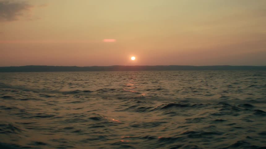 A wide shot shows the sun low in the sky as it sets over the dark ripples of a large body of water