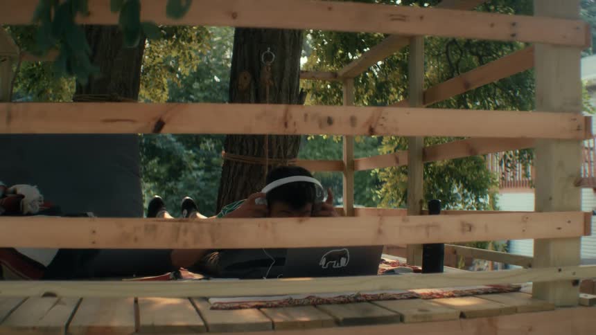 Person sitting in wooden treehouse putting on headphones and opening a laptop in his tree house to listen to music and do schoolwork