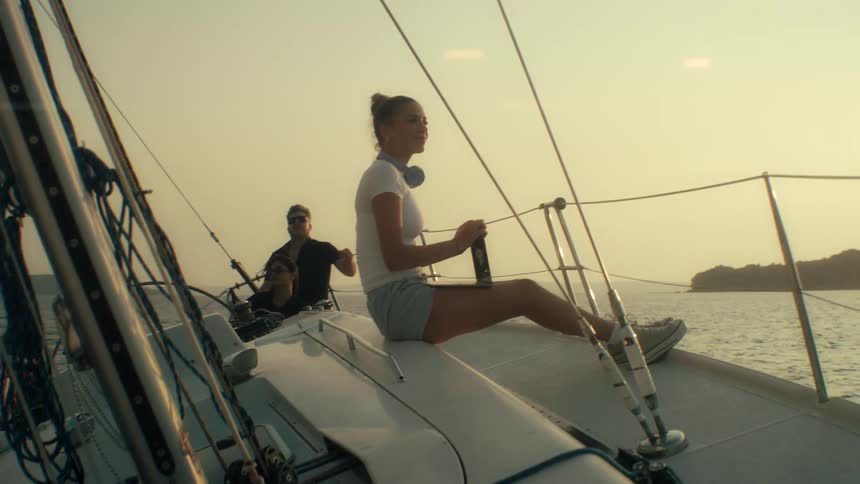 A young woman sits on the deck of a sailboat at sunset closing her laptop and looking out at the ocean while two men manage the vessel in the background near a distant coastline