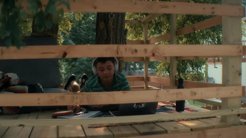 Teenager boy smiling while wearing headphones and writing on a notebook in front of a laptop computer lying down on the floor of a tree house during day time doing homework outside