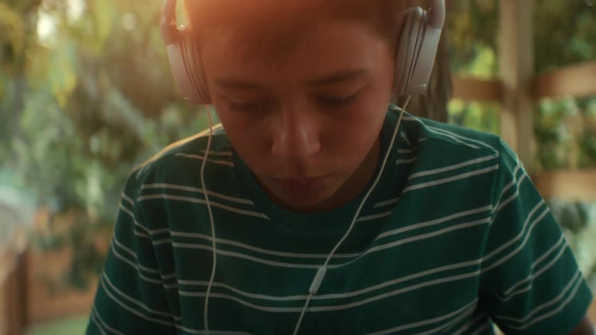 The boy is sitting under a tree with headphones on listening to music or an important sporting event or broadcast and typing