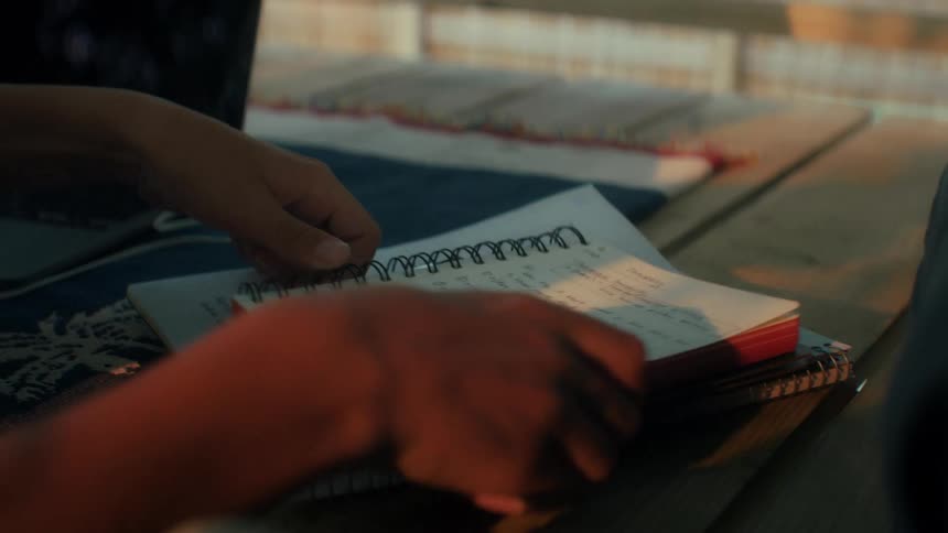 Someone doing homework outside is typing on their laptop and taking notes with a pen in the notebook in warm orange sunset light