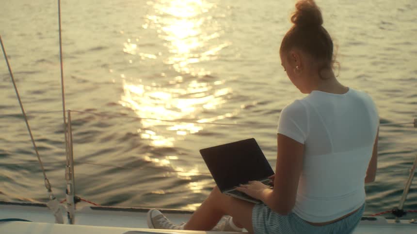 Person sitting on boat edge with laptop on lap at sunset over calm waters