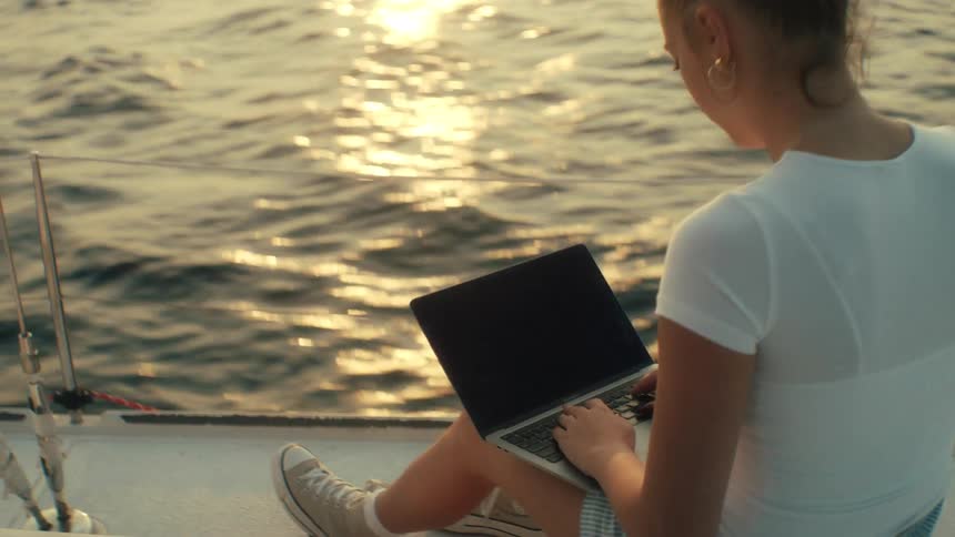A shot from behind shows a young woman sitting on a sailboat at sunset using a laptop while the sun reflects off the water