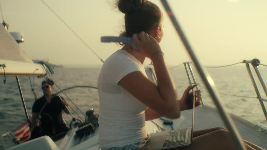 Woman in white shirt and striped shorts typing on laptop on sailboat deck at sunset with another person in background