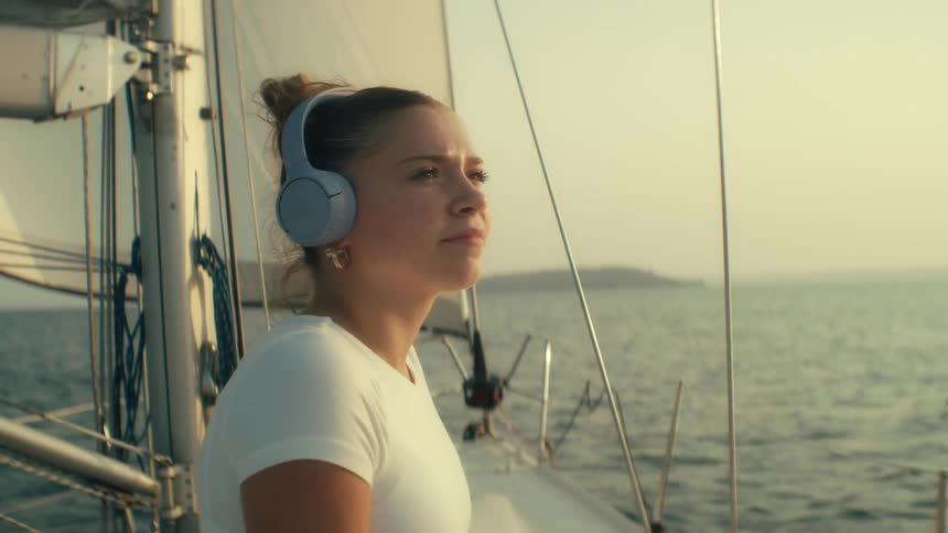 A woman with her hair in a bun wears blue headphones and a white shirt on a sailboat looking forward with a calm and contemplative mood