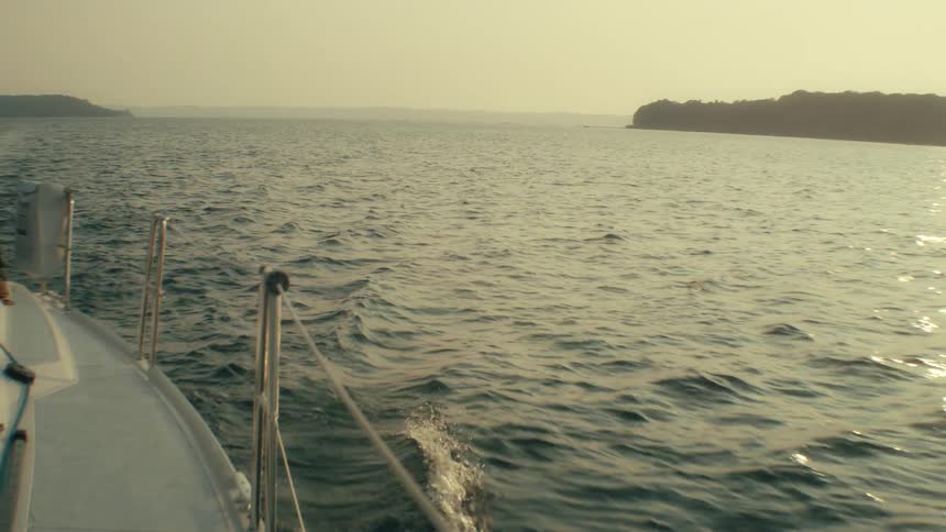A boat sails through choppy ocean waters toward a distant hilly coastline during a hazy sunset with the white deck and metal railings visible in the foreground as the vessel moves across the waves