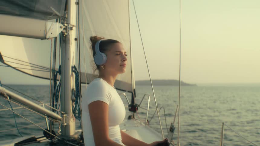 Teenager girl wearing headphones typing on a laptop computer then stopping and looking around the sea on a sailboat outdoors