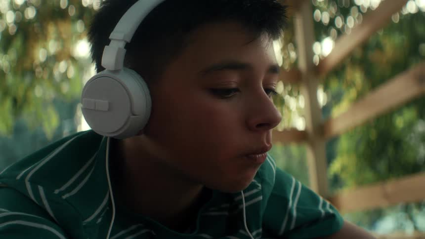 In a closeup a young boy with headphones on gazes downwards completely absorbed in what hes listening to while soft dappled sunlight filters through the trees behind him