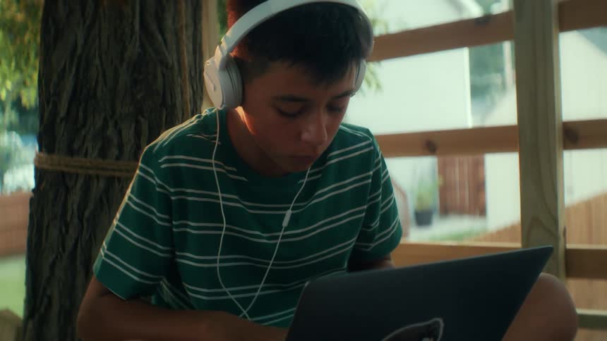 Teenage boy wears headphones as he sits outside listening to music focusing and typing on a laptop computer