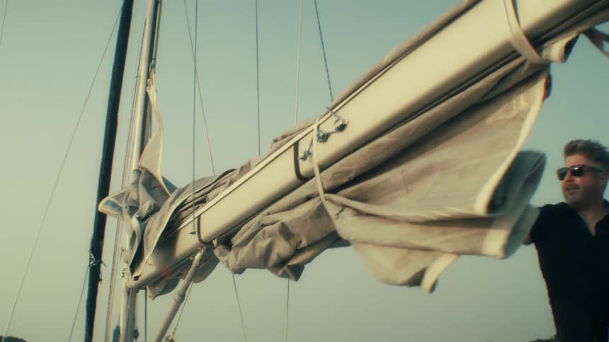 A man in a black polo shirt and sunglasses works on the deck of a sailboat pulling on ropes to release and unfurl a large grey sail from the boom against a clear sky during the day