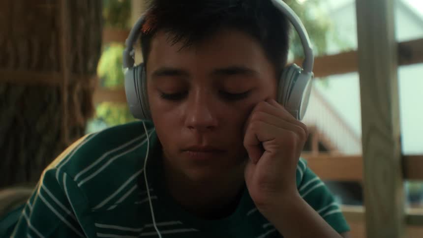 A boy laying in a tree fort puts on headphones to listen to music while homeschooling and doing homework