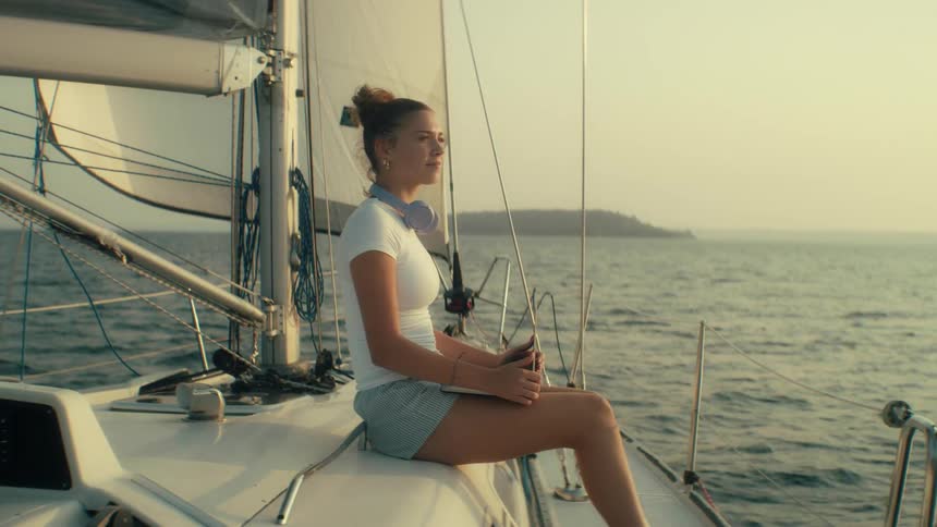 A medium shot shows a young woman sitting on the deck of a sailboat using a laptop and wearing headphones while looking out at the water as the sun sets