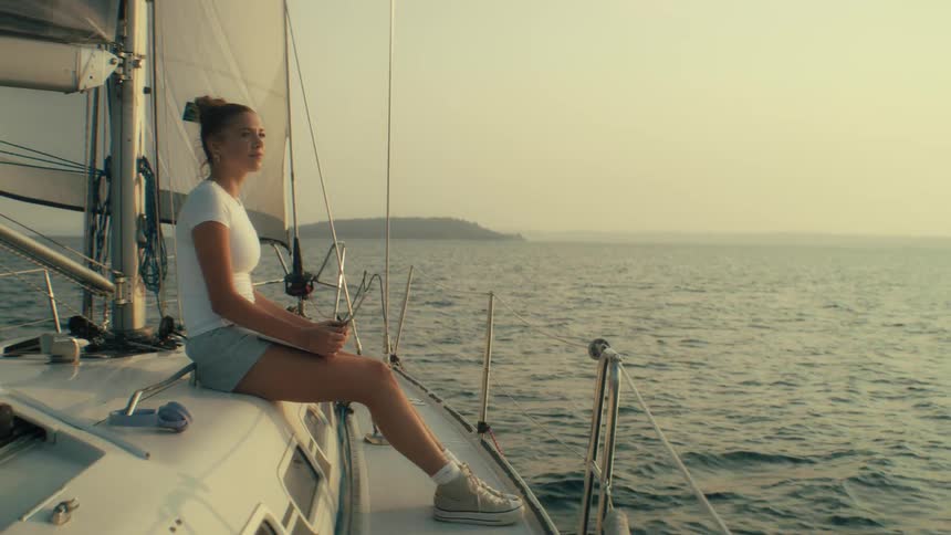 A medium shot shows a young woman sitting on the deck of a sailboat looking out at the water as the sun sets