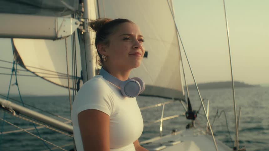 Teenager girl sitting and typing on a laptop computer then removing her headphones and looking around with a happy facial expression on a sailboat over sea waters outdoors during dusk or dawn