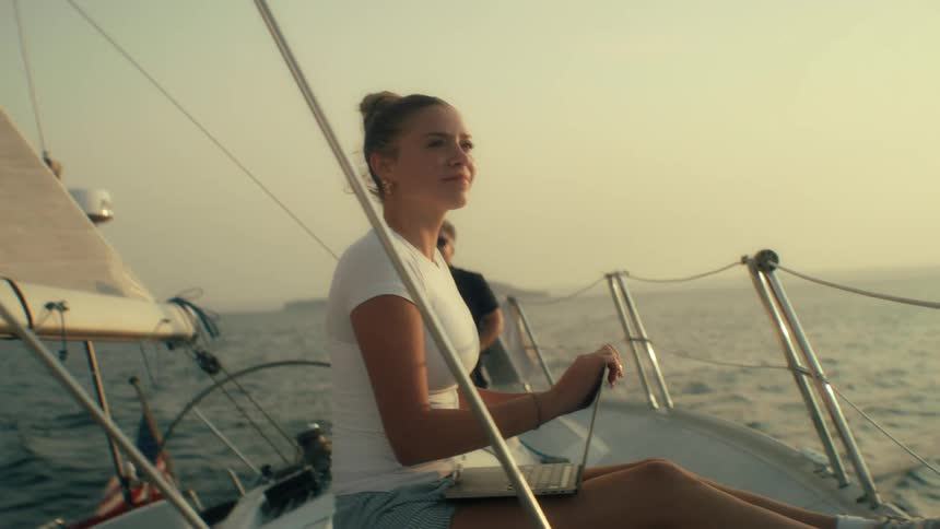 Young woman sits on a boat sailing off the coast as she holds a laptop computer with two other people on the deck