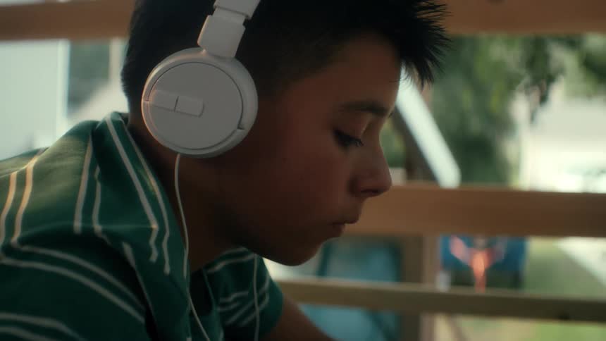Boy wearing headphones sitting outdoors in a treefort is thinking, listening, and typing