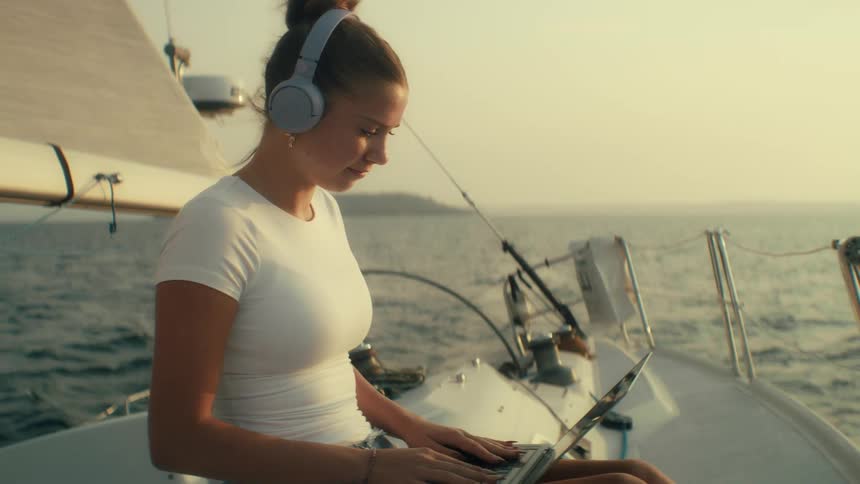 Young woman wearing headphones sits on a boat sailing off the coast and opens a laptop computer then types on the keyboard