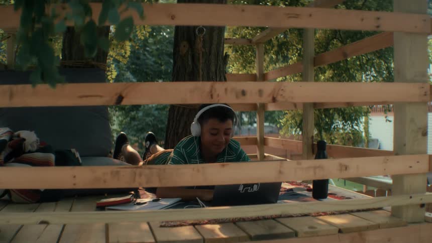 A kid lies on an unfinished tree house in the woods at sunset wearing a wired headset while using his laptop as warm light filters through the trees
