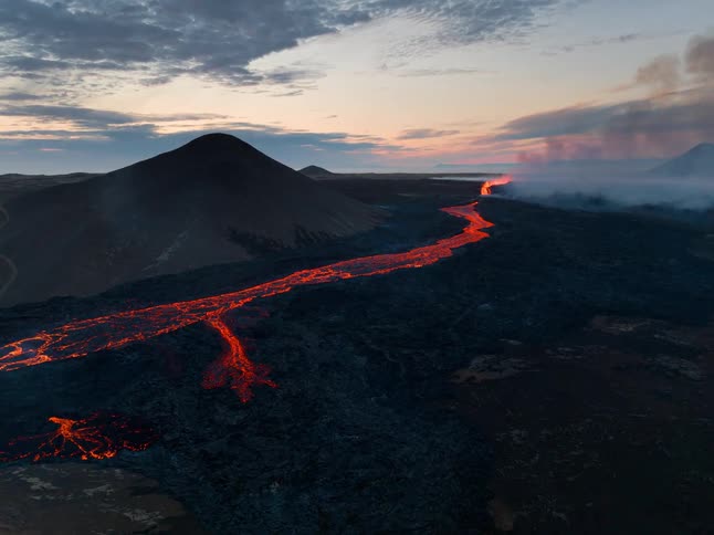 A wide aerial shot captures a bright river of molten lava flowing through a dark desolate volcanic field at dusk