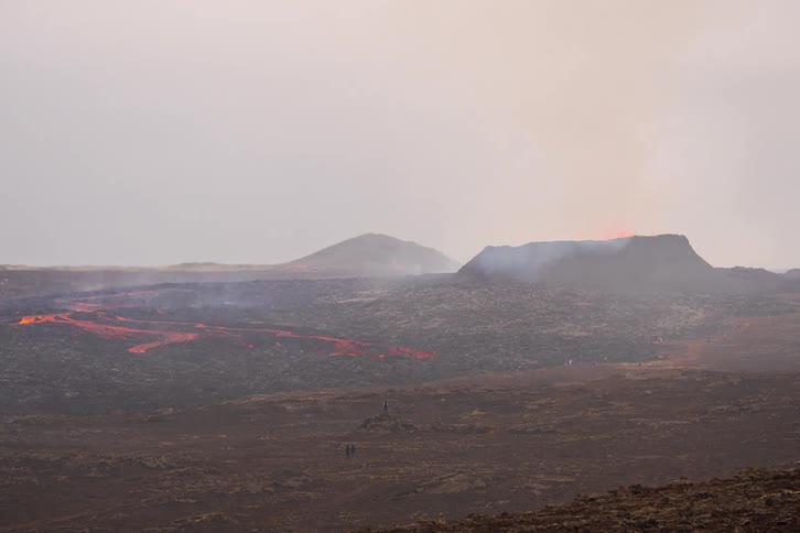 Smoke fills the air as people stand on high ground watching hot lava from an erupting volcano move across the land