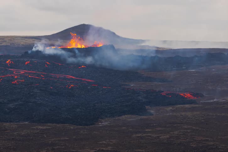 Smoke and flames emerge from an erupting volcano as molten hot lava moves across the land and people walk on high ground