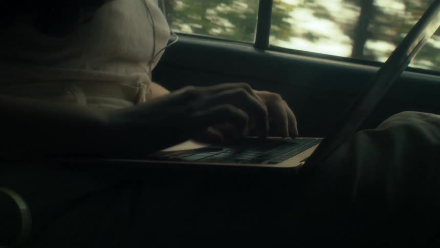 Teenage girl typing on the keyboard of a laptop computer as she sits in the back seat of an automobile