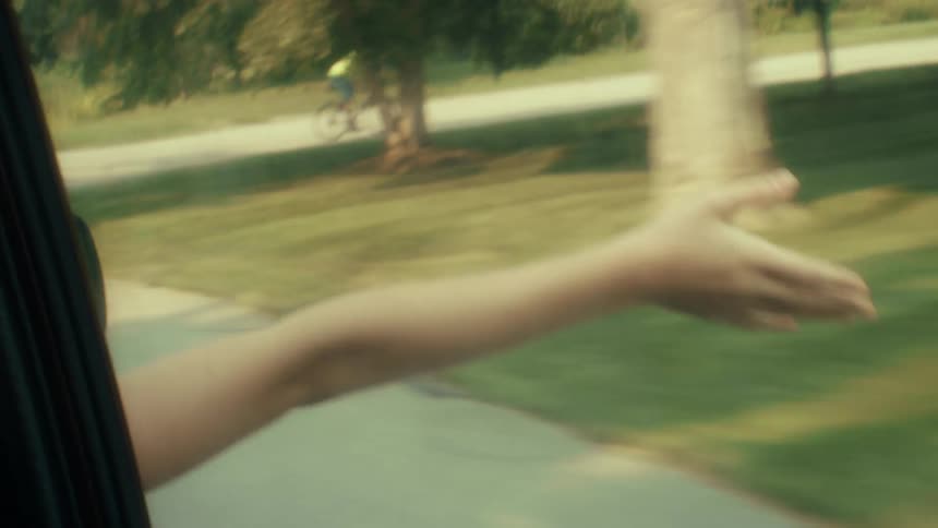 A person stretches a bare arm out of a moving car window toward a green field looking forward with a carefree mood