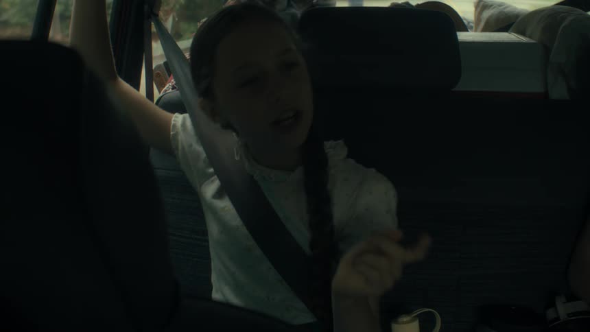 Inside a moving vehicle a young girl with a long braid wearing a white floral blouse sits in the back seat smiling and talking animatedly