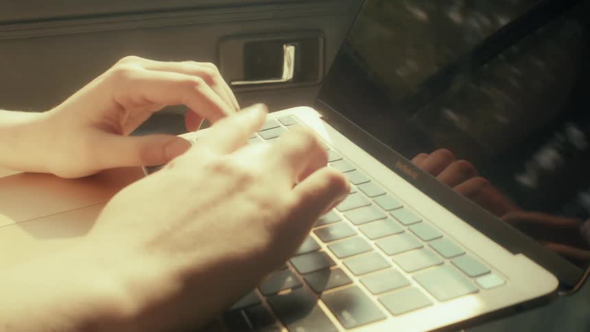 A woman sits inside a vehicle and types on a laptop keyboard with her hands moving quickly across the keys while the car door and window reflections are visible in the background